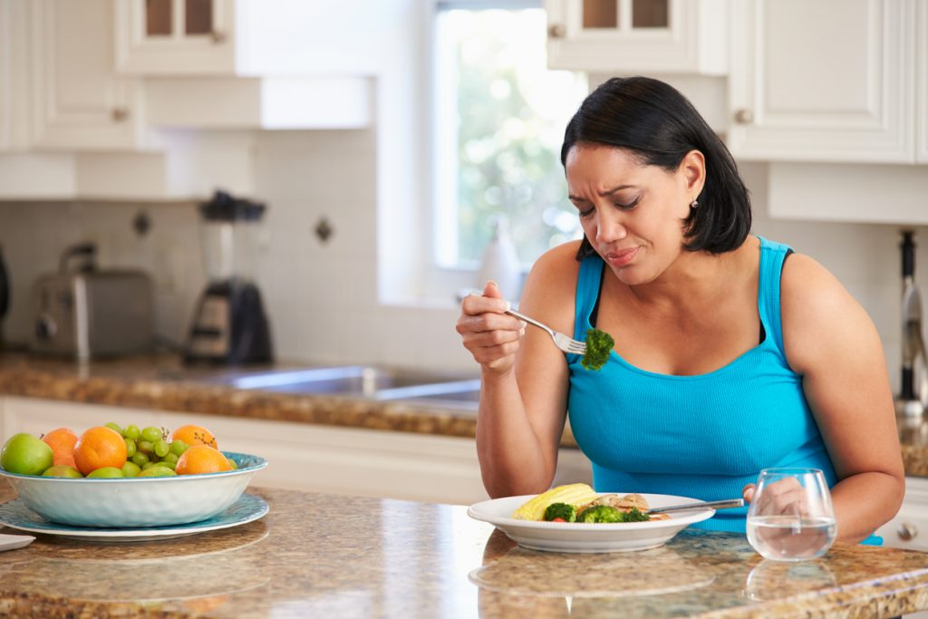 woman sitting at kitchen island with plate of food, looking dejectedly at broccoli on fork
